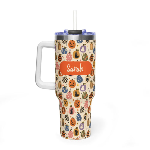 A handled insulated tumbler with a Halloween-themed pattern of pumpkins, ghosts, and bags of tricks on a cream background. The tumbler has a custom orange label with the name 'Sarah' written on it, a clear lid, and a white handle.