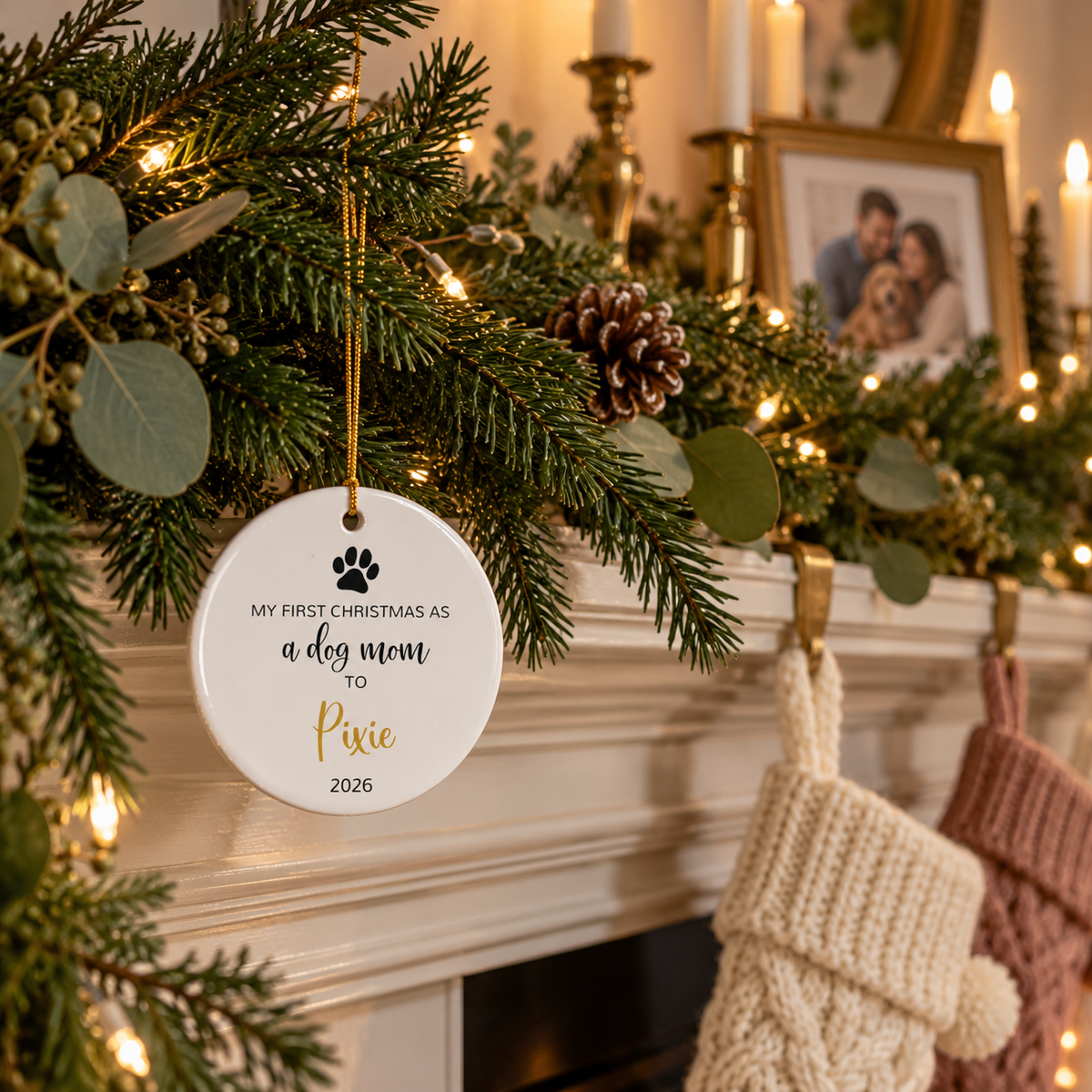 Personalized dog mom first Christmas ornament with paw print and gold script name on a fireplace garland with stockings